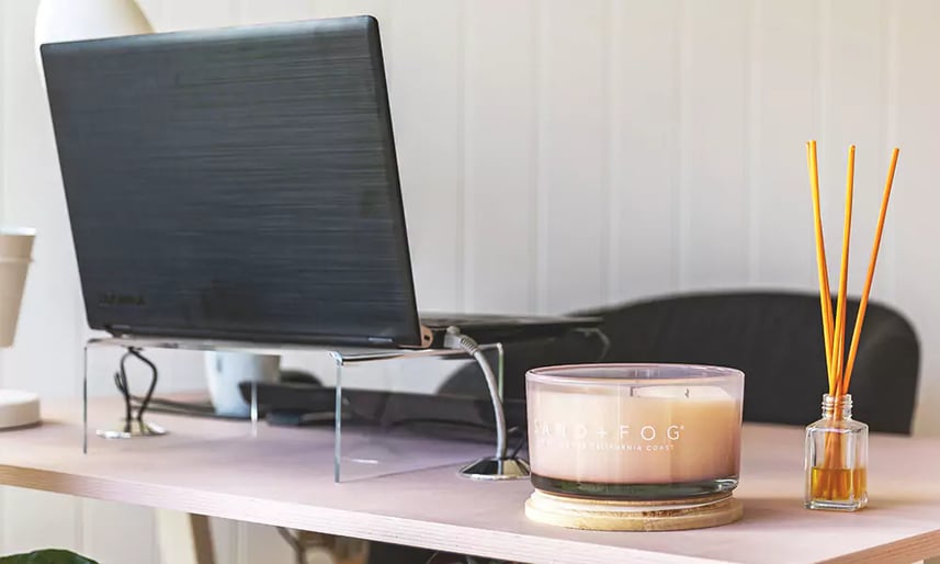 A light wooden desk with a small reed diffuser, a pink candle and a laptop. The laptop is sat on a glass stand and there is a keyboard on the desk behind it. In the background there is a grey office chair and a white, wood panelled wall.