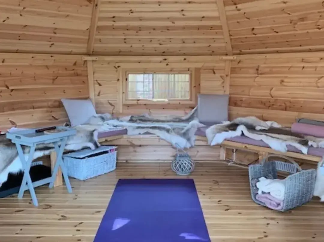 A small hexagonal log cabin with a purple yoga mat in the centre. The benches around the edge of the cabin have cushions and reindeer skins around dotted around.