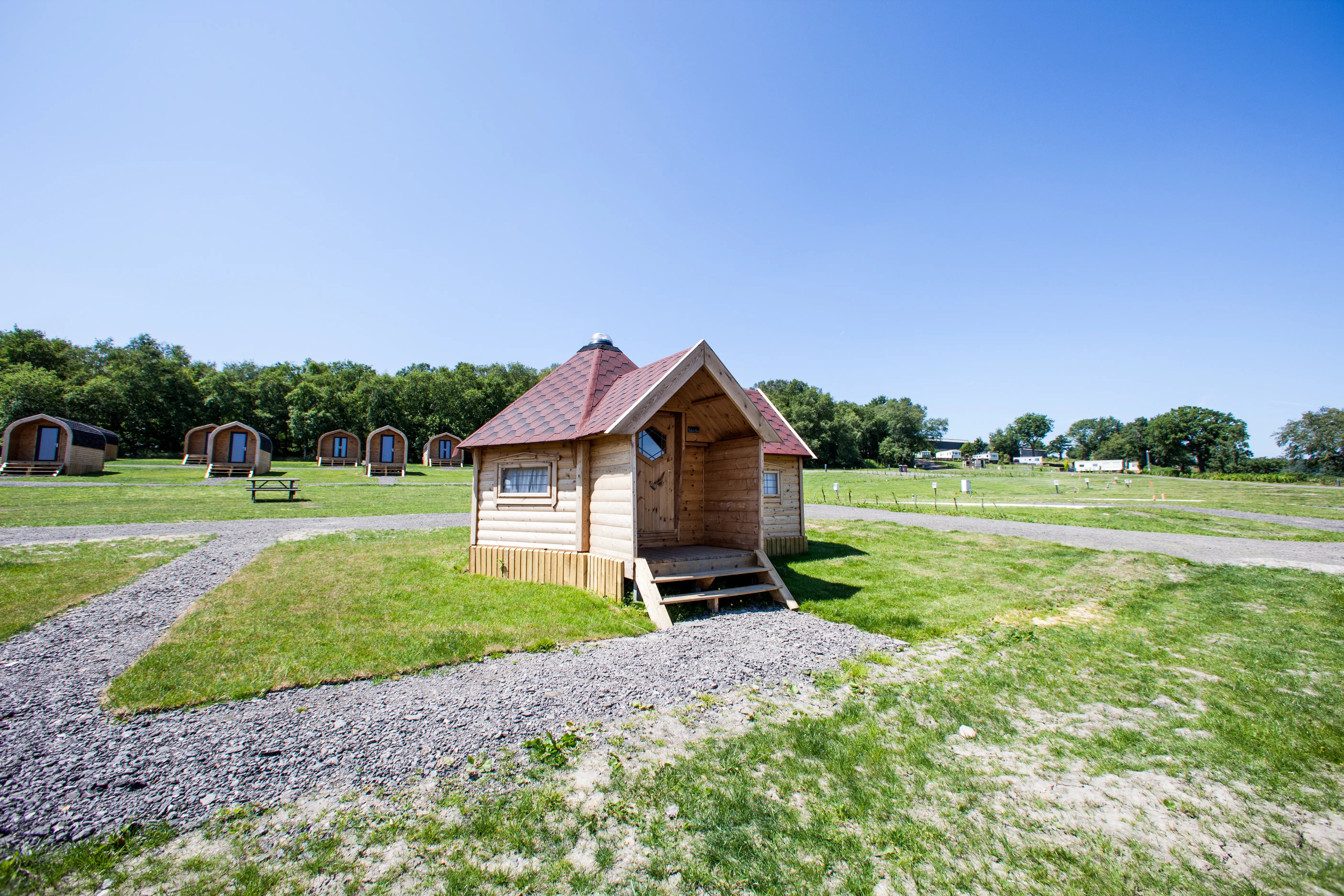 An extended hexagonal glamping log cabin with a sheltered porch. The cabin has a peaked red shingle roof and is sat in a grass field with a straight gravel path leading up to the porch. Further behind the cabin there are arched glamping pods in a row.