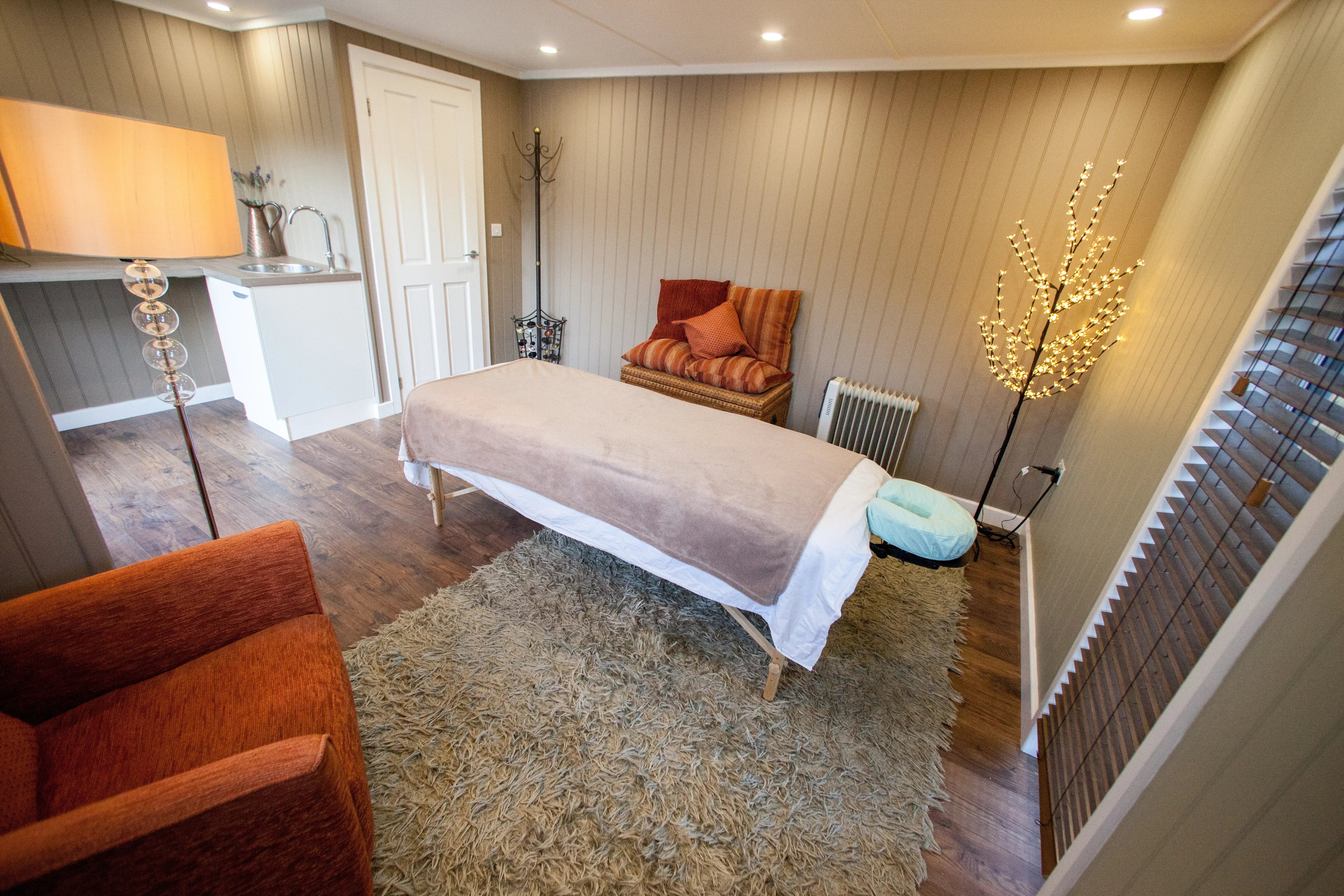 Modern rectangular wellness cabin with a massage cabin in the centre. The are floor lamps in each opposite corners of the cabin and a desk and sink unit near the entrance.