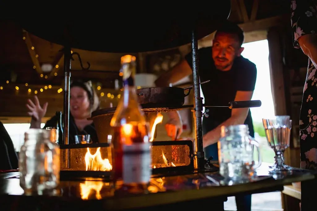 A close up of the central BBQ grill in a cabin. There are drinks and bottles around the table and a man is cooking food on the grill while guests are sat chatting.