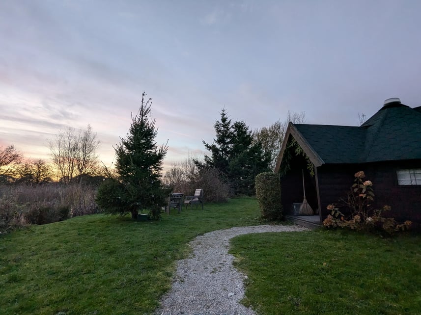 Driftwood Lodges glamping cabin at sunset. There is a set of wooden chairs and a table next to an evergreen tree and a gravel path leading up to the porch of the cabin.