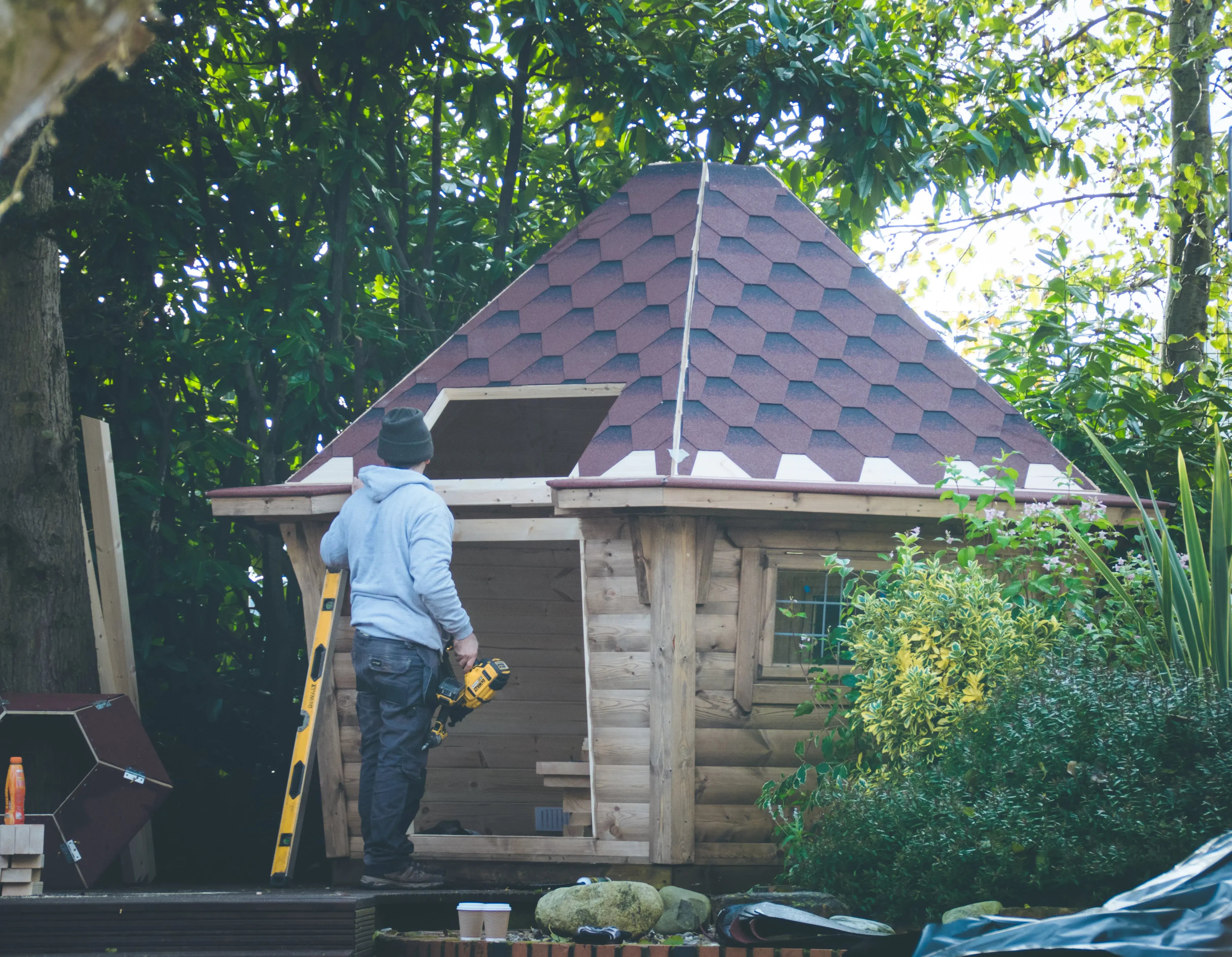 A man in a light grey hoodie, black trousers and dark grey beanie, holding a yellow and black drill in front of a BBQ log cabin. The BBQ cabin is being built and doesn't have the door or porch attached. To the right side of the cabin there are various green bushes and trees behind the BBQ hut.