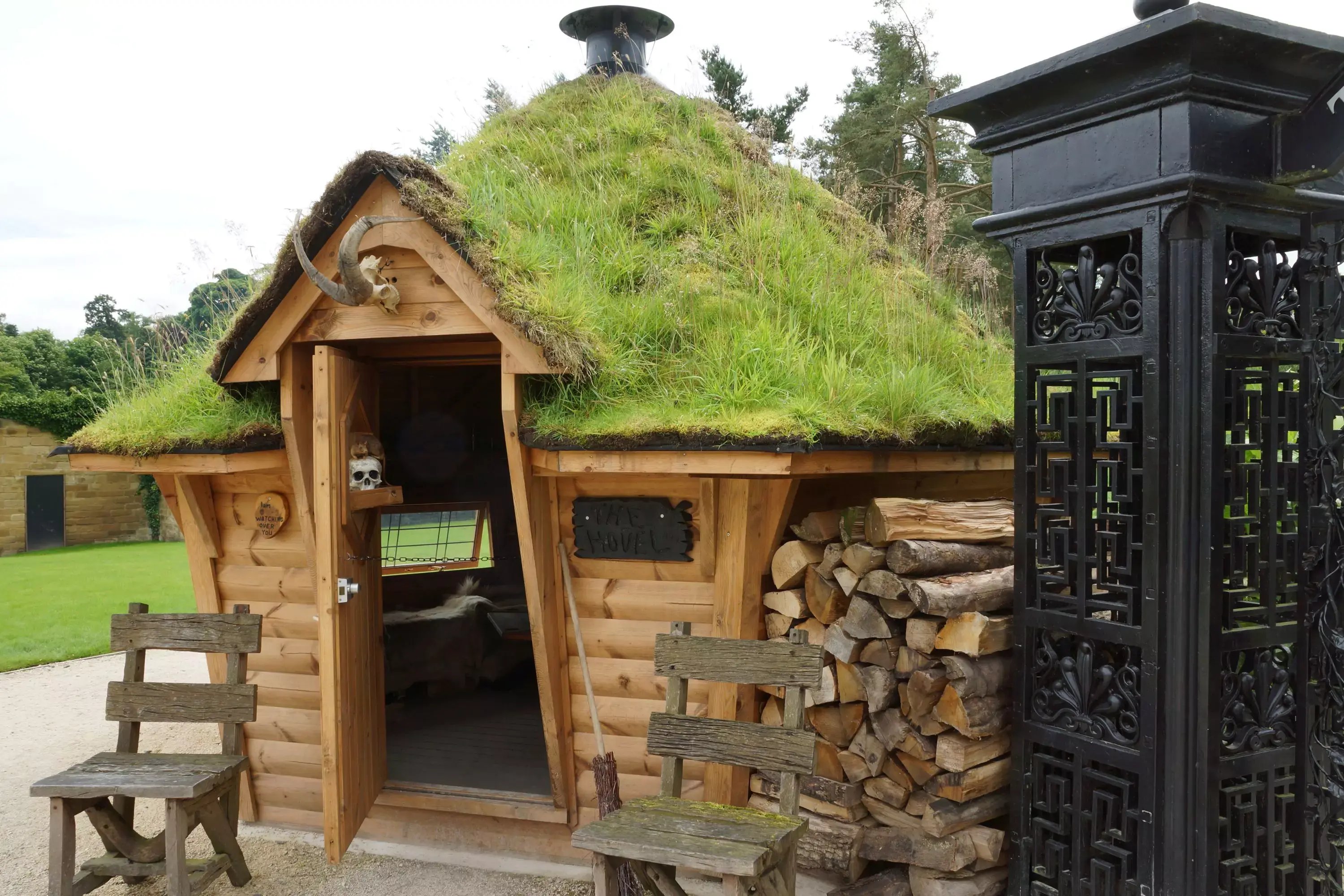 Witches log cabin at Alnwick Castle. The log cabin has a grass roof and the door is decorated with old skulls. There is a neatly stacked pile of firewood to the right of the cabin.