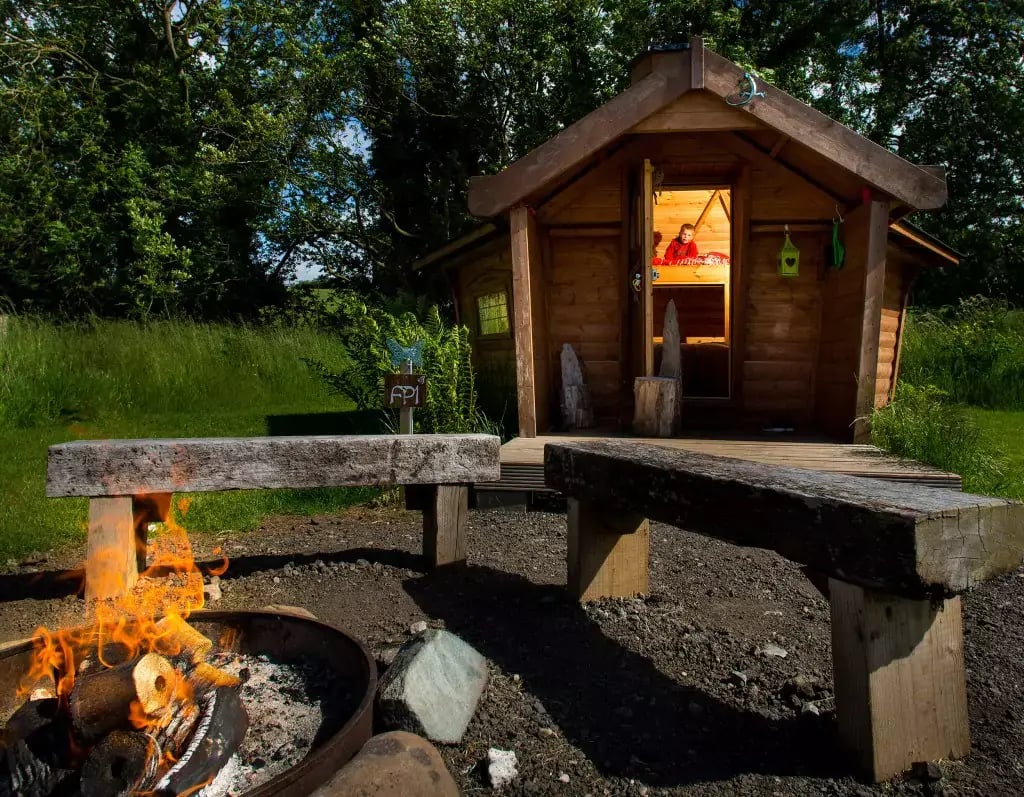 A firepit in front of a wooden glamping pod. There are two wooden benches next to the lit campfire. In the glamping cabin, there is a young child sat on the top bunkbed, looking outside. Behind the glamping cabin there is green grass, bushes and trees.
