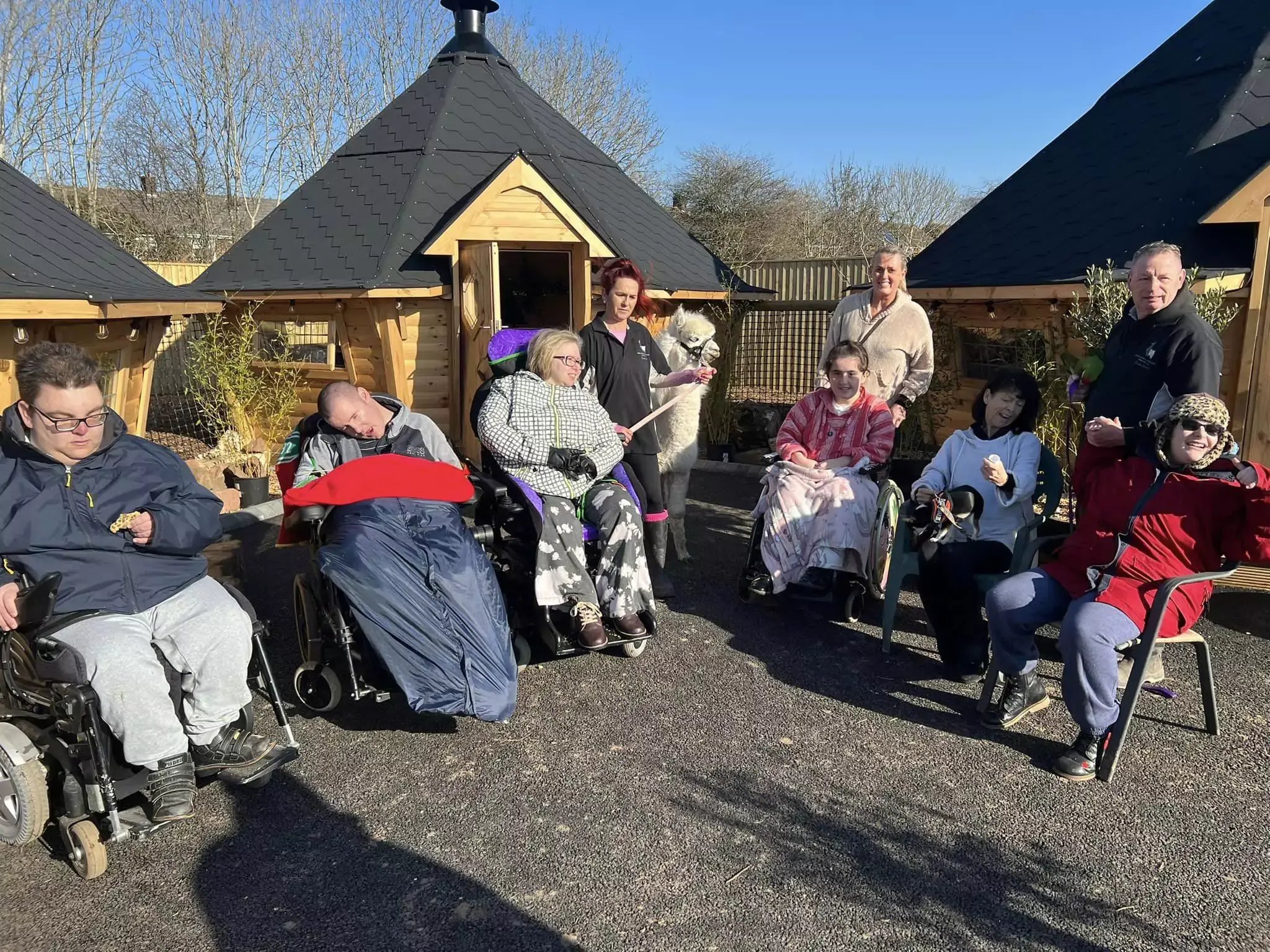 A group of people in wheelchair and mobility aids and their carers outside 3 BBQ huts at Nanny Sharon's Alpaca Farm. In the middle there is a woman with red hair leading an alpaca.