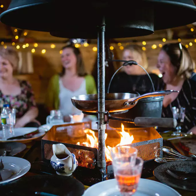 A group of women are sat around a lit central BBQ grill in a log cabin. There are plates and glasses on the table surrounding the grill.