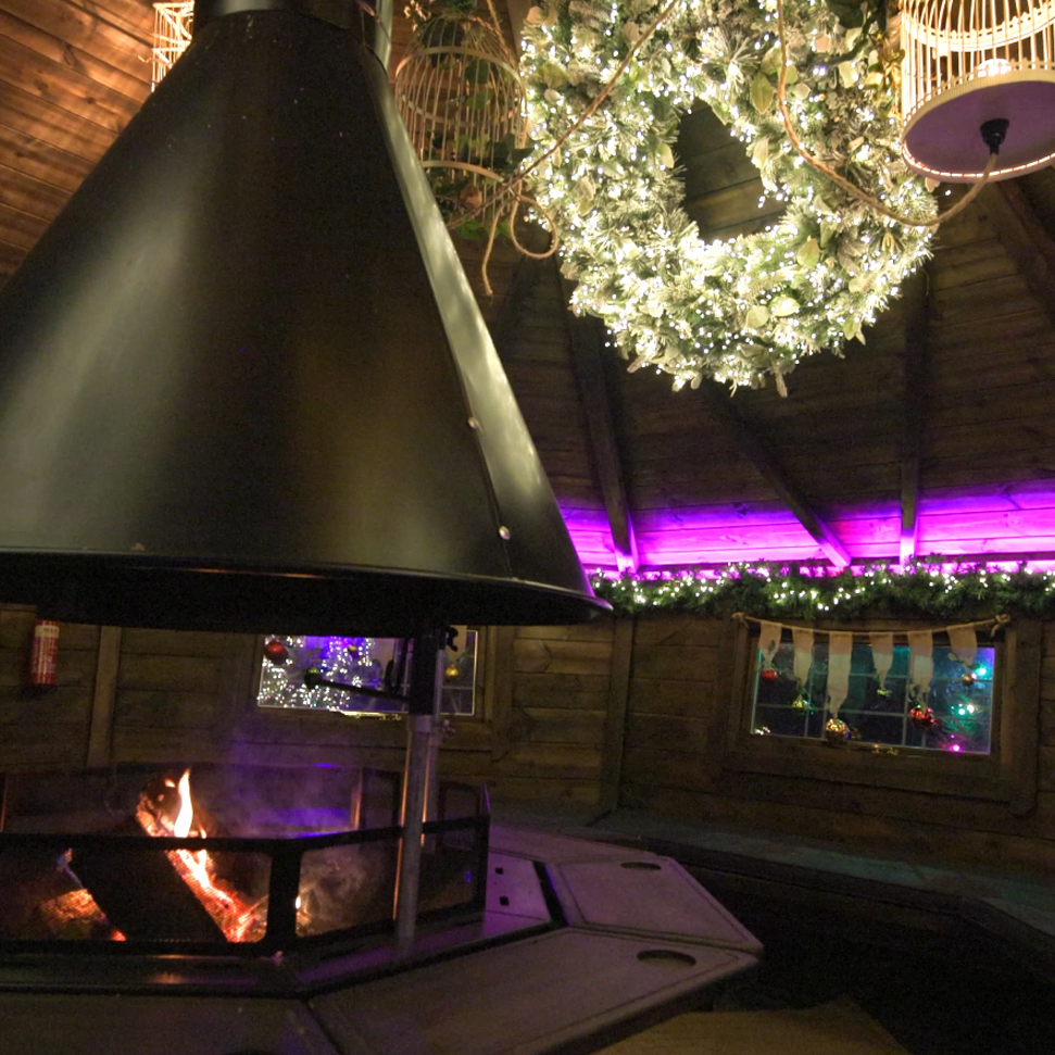 The interior of a BBQ cabin with a large Christmas wreath and decorative bird cages hanging from the roof. There is an evergreen garland with fairy lights lining the wall of the cabin and baubles hanging from ribbons above the windows.