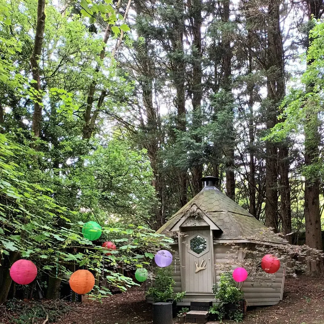 Exterior of a BBQ log cabin that has been painted a light grey. The cabin is surrounded by tall trees and there are colourful paper lanterns hanging from the tree in front of the cabin.