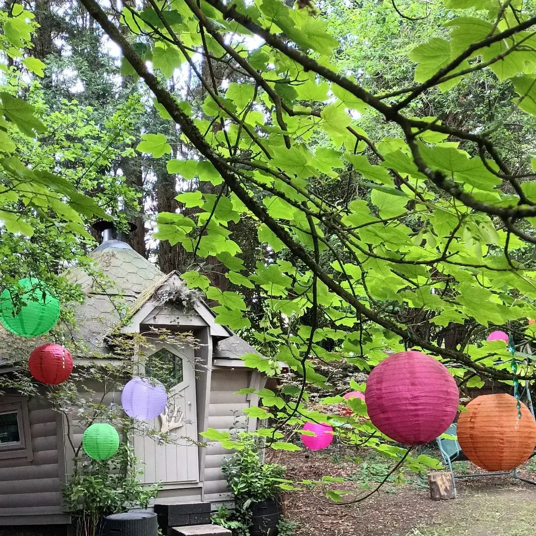 Exterior of a BBQ log cabin that has been painted a light grey. The cabin is surrounded by woodland and there are colourful paper lanterns hanging from the tree in the foreground.
