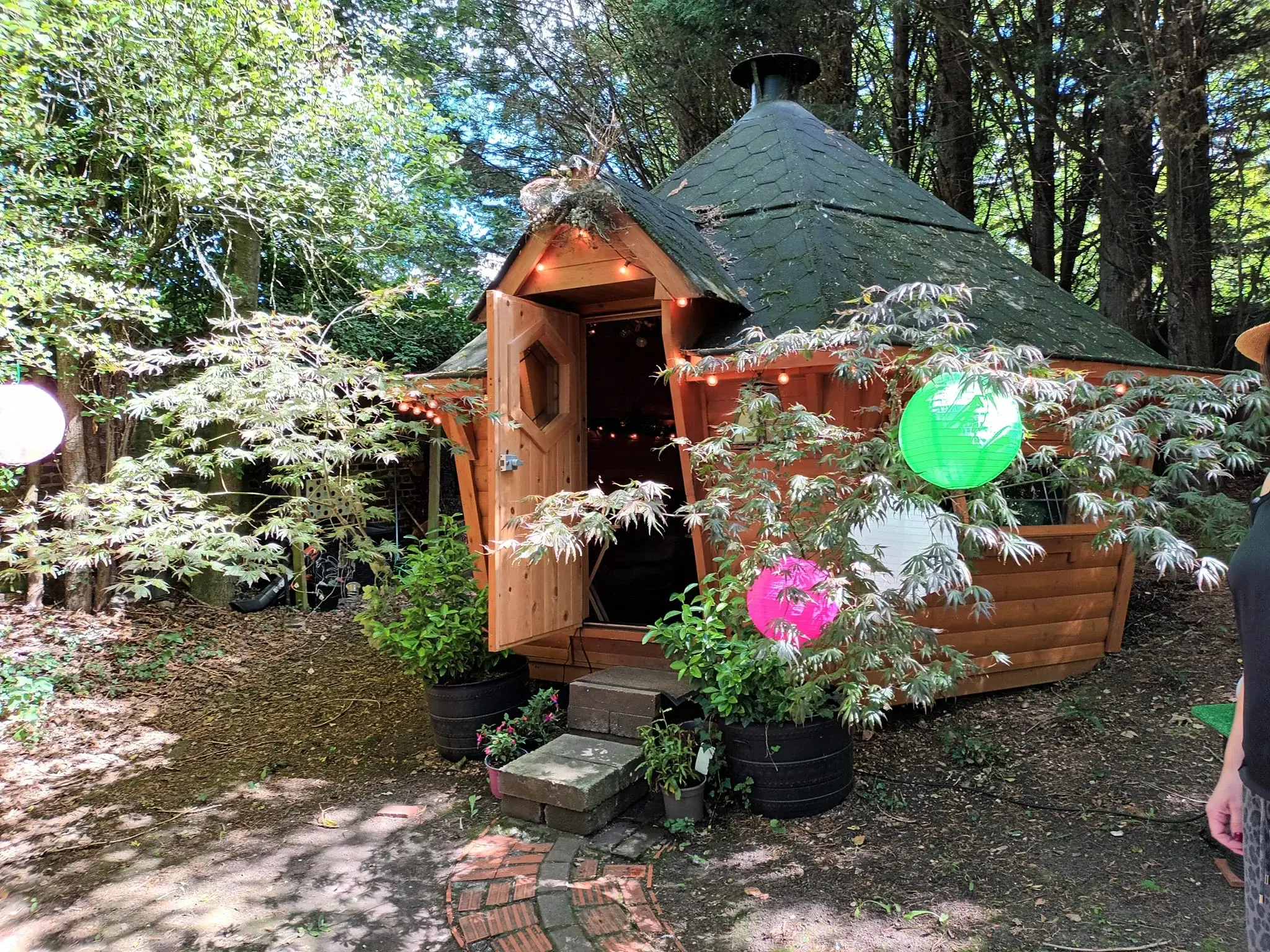 A hexagonal log cabin with a peaked green roof in a woodland surrounding. There are colourful paper lanterns in the trees surrounding the cabin.